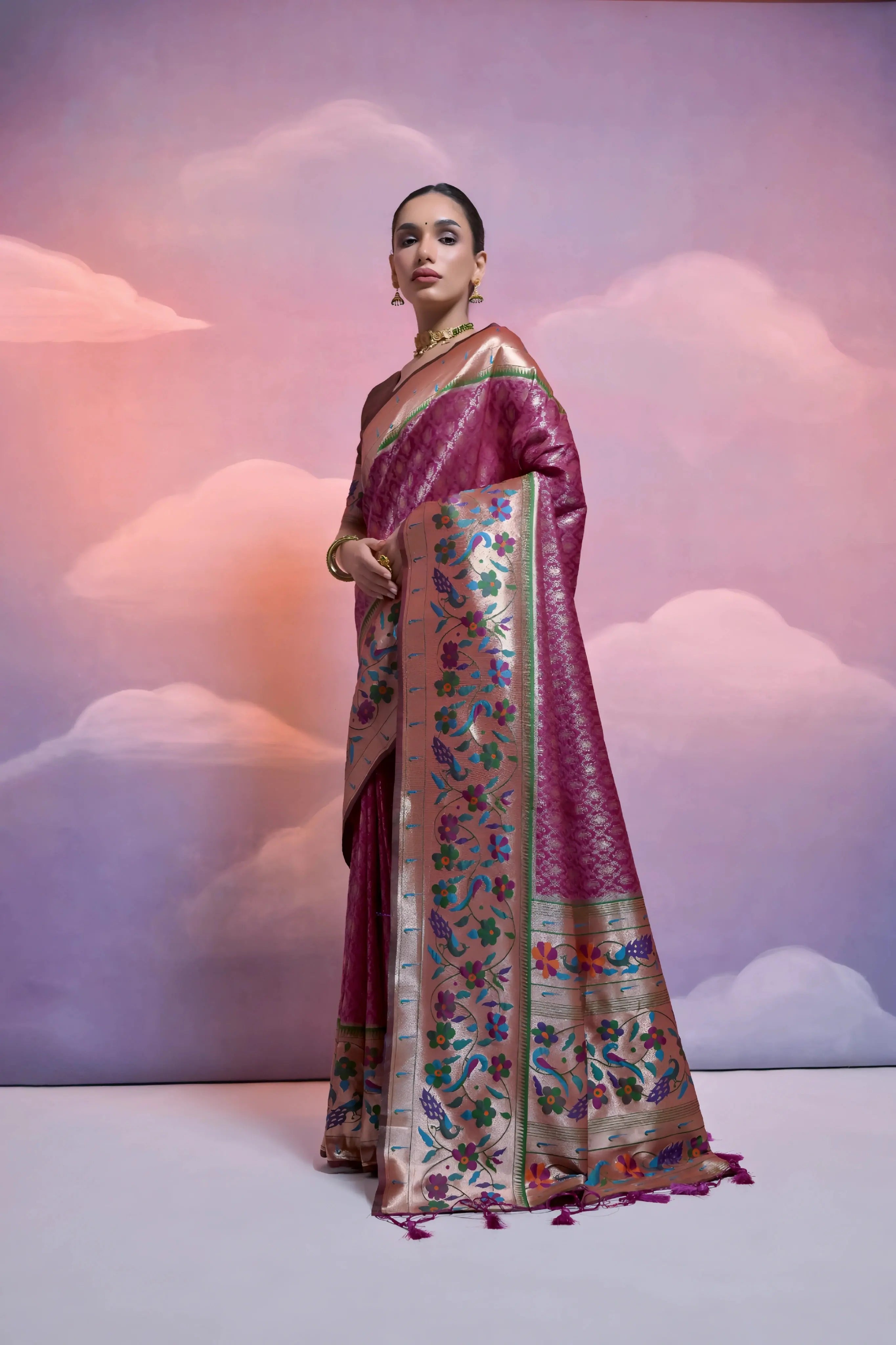A woman models a luxurious Wine Red Soft Silk Saree with dense all-over weaving. The wide border and pallu feature heavy, multi-colored floral and figurative weaving in gold, green, blue, and pink threads. She wears a dual-toned blouse and heavy gold jewelry with green gemstone accents, posing against a scenic painted backdrop.